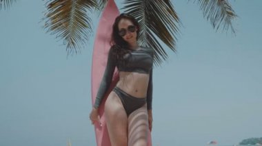 Güzel genç kız yaz tatilinde / güzel surfboard ile genç kadında mayo ve palmiye ağacı ve gökyüzü arka plan - ağır çekimde video üzerinden güneşli yaz gün boyunca pembe surfboard ile poz güneş gözlüğü