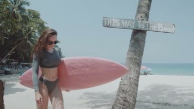 Güzel genç kız yaz tatilinde surfboard ile / mayo ve güneş gözlüğü mutlu genç kadında palmiye ağacı ve gökyüzü arka plan - ağır çekimde video üzerinden güneşli yaz gün boyunca pembe surfboard ile yürüyüş