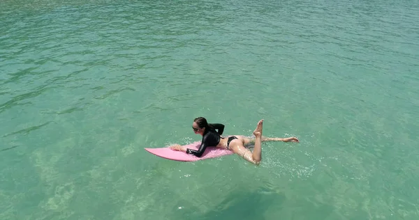 水着とサングラスに驚くほど澄んだ海の水で彼女の休日を楽しんでいるピンクのサーフボードの美しい若い女性の空中ドローン ビュー — ストック写真