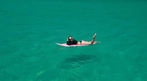 水着とサングラスに驚くほど澄んだ海の水で彼女の休日を楽しんでいるピンクのサーフボードの美しい若い女性の空中ドローン ビュー — ストック写真
