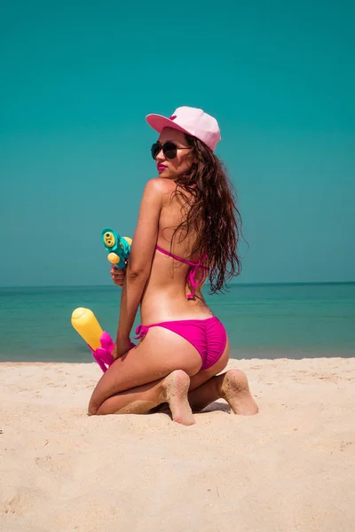 ピンクのビキニ キャップとブルーのサングラスが海と空の背景に砂浜に水銃を持ったポーズで笑顔美人 — ストック写真