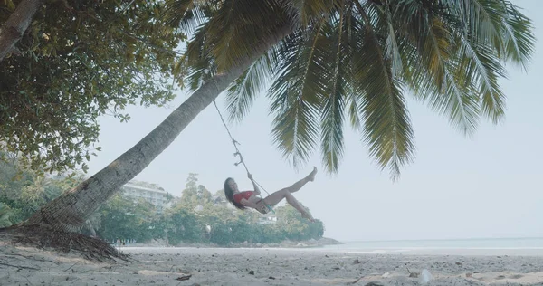 水着で熱帯のビーチでヤシの木にぶら下がっているブランコに乗っているリラックスした美しい幸せな少女 — ストック写真