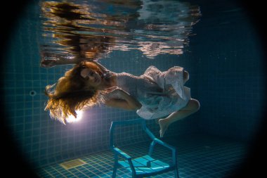 Uzun kızıl saçlı, beyaz elbiseli, sandalyenin altında poz veren güzel bir kadın.