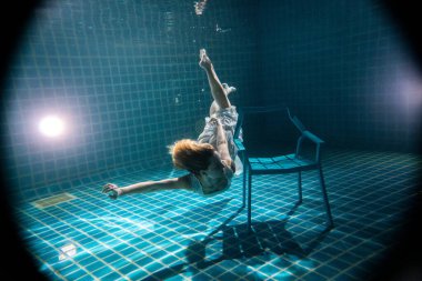 Suyun altında beyaz elbiseli sandalyenin yanında poz veren uzun kızıl saçlı güzel bir kadın.