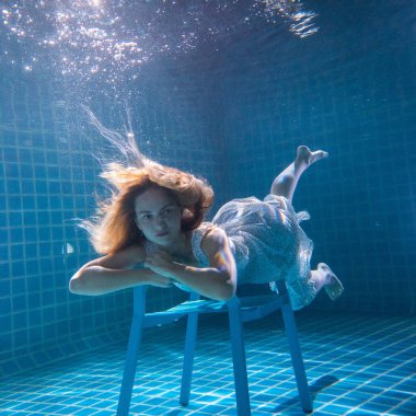 Uzun kızıl saçlı, beyaz elbiseli, sandalyenin altında poz veren güzel bir kadın.