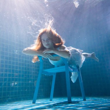 Uzun kızıl saçlı, beyaz elbiseli, sandalyenin altında poz veren güzel bir kadın.