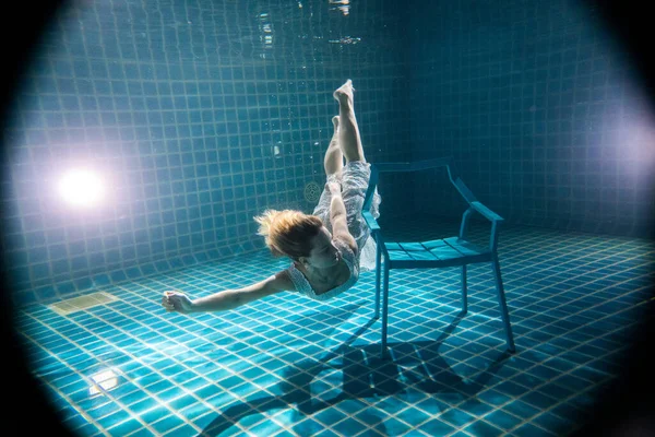 Suyun altında beyaz elbiseli sandalyenin yanında poz veren uzun kızıl saçlı güzel bir kadın.