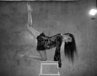 Beyaz dantelli iç çamaşırlarıyla güzel bir akrobatik. Stüdyonun ahşap sandalye merdiveni üzerinde poz veriyor. Duvarın arkasında.