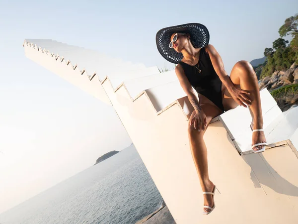 黒水着のファッション女の子 帽子とサングラスは空と海の背景の上の階段の上に屋外でポーズ — ストック写真