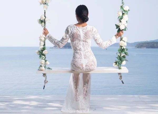白いレースのドレスで美しい女性のバックビューは 海と空の背景に花と屋外スイングにポーズ — ストック写真