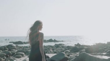 Siyah ipeksi elbiseli, yürüyen, koşan, yaz günü kumsalda kamerayla flört eden güzel esmer kadın.