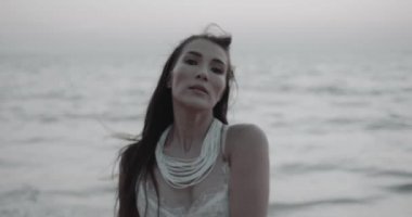 Beyaz dantel elbiseli güzel esmer kadının romantik portresi yaz akşamının erken saatlerinde kumsalda kamerayla flört ediyor. Yavaş çekimde video.