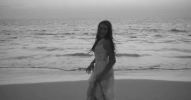 Beyaz dantelli elbisesi olan güzel esmer kadının romantik portresi yaz akşamının erken saatlerinde kumsalda kamerayla flört ediyor. Yavaş çekimde siyah beyaz bir video.