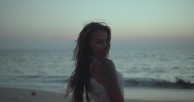 Beyaz dantel elbiseli güzel esmer kadının romantik portresi yaz akşamının erken saatlerinde kumsalda kamerayla flört ediyor. Yavaş çekimde video.