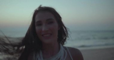 Beyaz dantel elbiseli güzel esmer kadının romantik portresi yaz akşamının erken saatlerinde kumsalda kamerayla flört ediyor. Yavaş çekimde video.