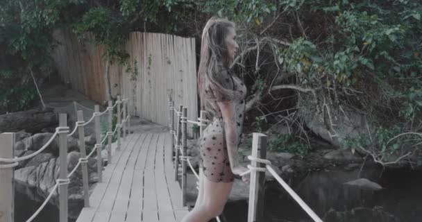 Gyönyörű barna nő séta, flörtölés a kamerával át kis fa híd a nyári nap folyamán - videó lassított felvételen