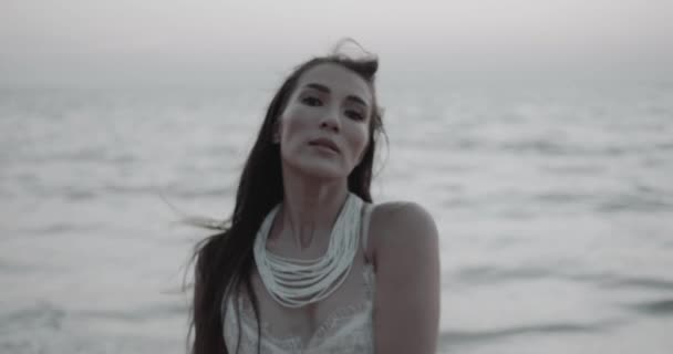 白いレースのドレスを歩くの美しいブルネットの女性のロマンチックな肖像画 夏の早朝の夜の間にビーチでカメラといちゃつく ビデオでスローモーション — ストック動画