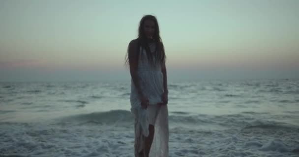 白いレースのドレスを歩くの美しいブルネットの女性のロマンチックな肖像画 夏の早朝の夜の間にビーチでカメラといちゃつく ビデオでスローモーション — ストック動画