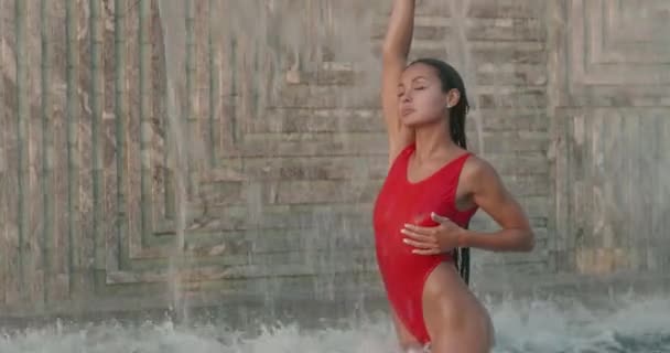 美しいブルネット女性で赤い水着姿で屋外プールの上に壁に滝の背景 ビデオでスローモーション — ストック動画