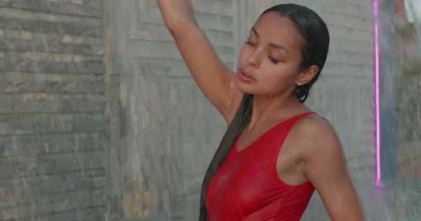身穿红色泳衣的漂亮的黑发女子站在户外游泳池里 背对着瀑布 慢镜头 — 图库视频影像