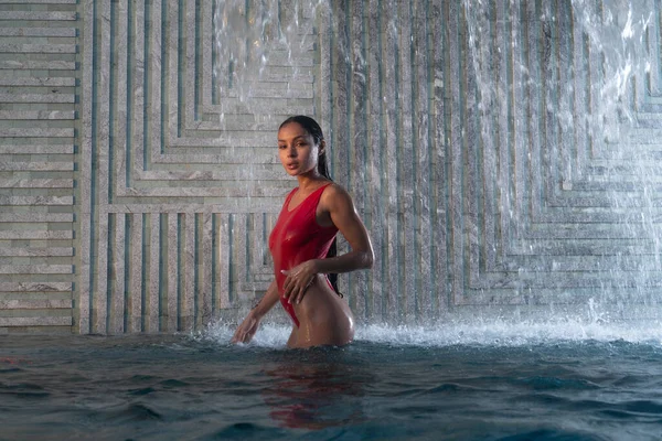 身穿红色泳衣的漂亮的黑发女人站在有瀑布背景的室外游泳池里 — 图库照片