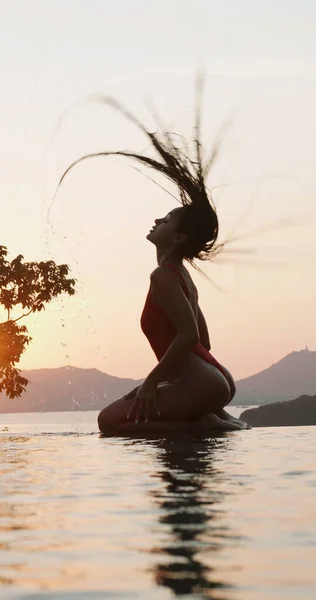 美丽的黑发女子 身穿红色泳衣 站在落日的天空背景下的室外游泳池前 — 图库照片