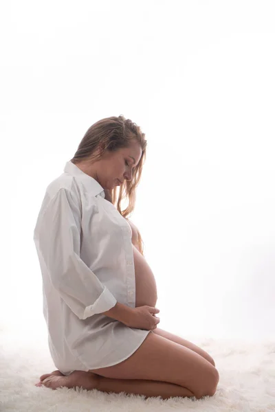 白い背景にひざまずきながらお腹を抱えている白いシャツとパンティーを着たかなり妊娠中の女性 産前概念 — ストック写真