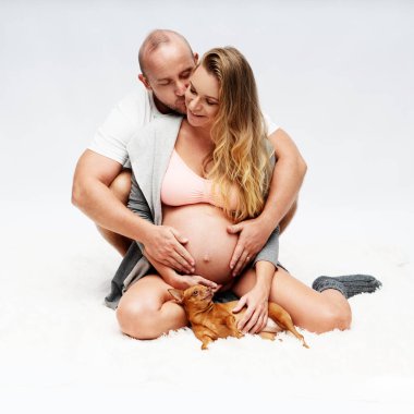 Sevgi dolu bir adam beyaz duvar arkasında sevimli küçük bir köpekle otururken hamile karısına arkadan sarılıyor. Annelik, hamilelik, yeni hayat ve aile planlaması kavramı