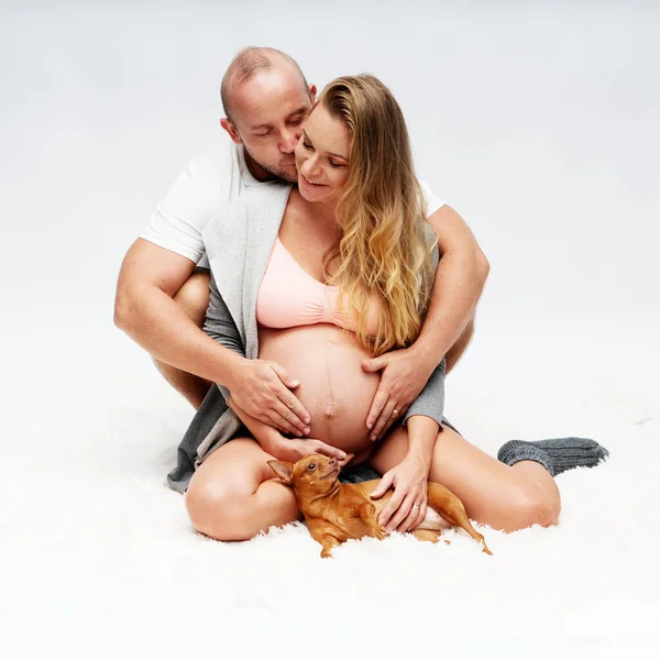 白い壁の背景にかわいい犬と一緒に座っている間に後ろから彼の妊娠中の妻を抱きしめる男を愛し 新しい生活と家族計画の概念 — ストック写真
