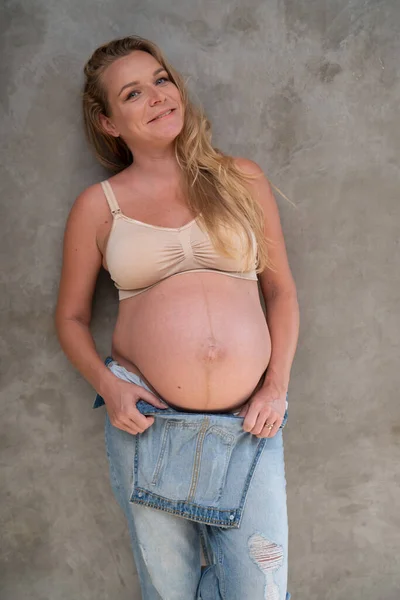 幸せなかなり妊娠中の女性デニムジャンプスーツとブラジャーグレーの壁の背景に立っている 産前概念 — ストック写真