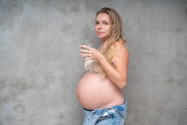 グレーの壁の背景の上に立っている間 デニムジャンプスーツや水のブラジャー保持ガラスでかなり妊娠中の女性 新しい生命の健康食品の概念 — ストック写真