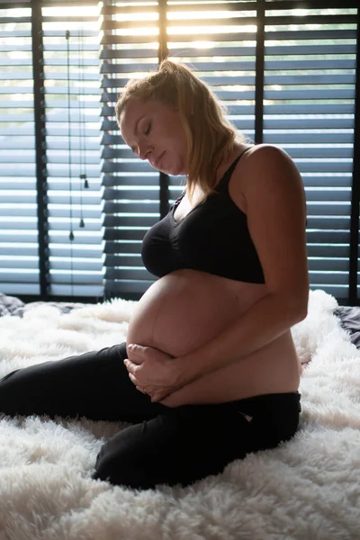 黒のレギンスとブラジャーのかなり妊娠中の女性は 自宅で彼女のベッドに座っている間に腰痛に苦しんでいます 妊産婦 健康管理 婦人科 — ストック写真