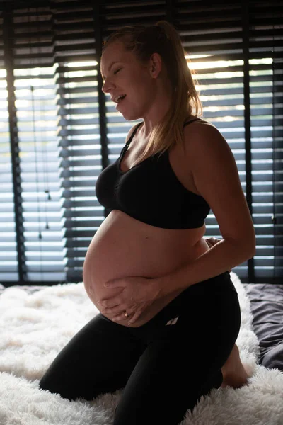黒のレギンスとブラジャーのかなり妊娠中の女性は 自宅で彼女のベッドに座っている間に腰痛に苦しんでいます 妊産婦 健康管理 婦人科 — ストック写真