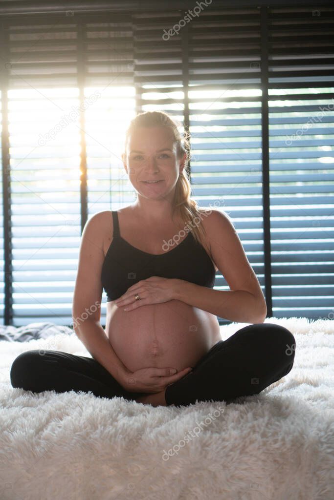 Happy pregnant woman in black leggins and bra holding her belly while relaxing on her bed at home. Maternity concept