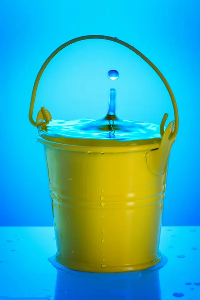 A small yellow bucket with water on a blue background. A drop of water falls into it and creates a splash of water.