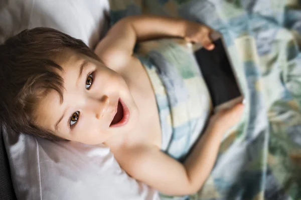 Garoto menino feliz segurando telefone celular deitado na cama de manhã — Fotografia de Stock