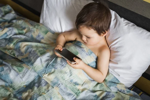 Moderna pojke spelar telefonen ligger på sängen i morgon — Stockfoto
