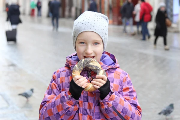 La fille mange un bagel avec du pavot dans une rue de la ville, jour froid . — Photo