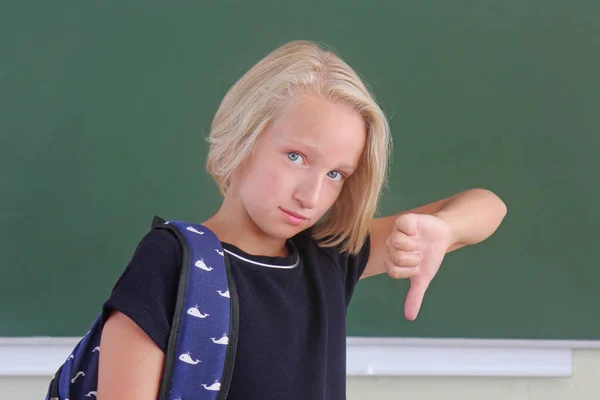 Sad schoolgirl with a backpack is showing a thumbs down in a classroom near green chalkboard. The child does not like the school, girl shows a gesture of a dislike. Back to school. — Stock Photo, Image