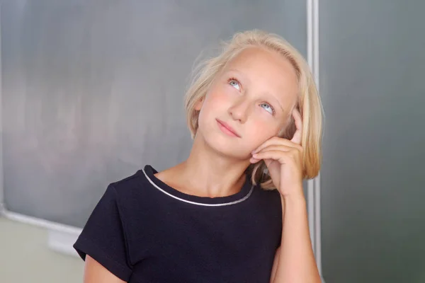 Adorable blond kid girl thoughtful in a classroom near a chalkboard. The child remembers, looking up thoughtfully. Back to school. — Stock Photo, Image