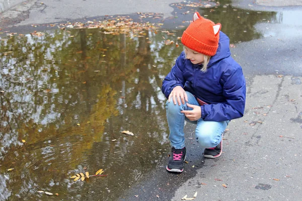 Kid girl in a warm knitted cap playing with leaves near a puddle while walking in an autumn park. — Stock Photo, Image