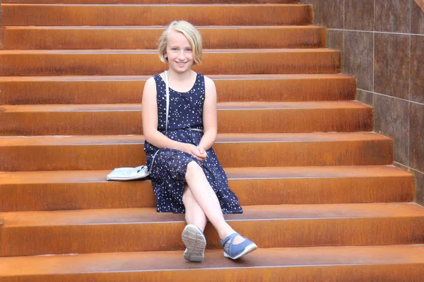 Stylish blonde preteen girl in a thin dress sits on the stairs with decorative rust in a modern building