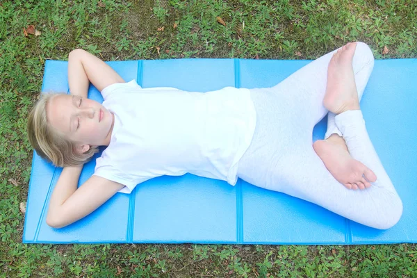 Okul çağındaki kız hafif giysiler içinde bir dinlenme ve spor, gözleri kapalı çalış, parkta mindere yalan söylüyor. Yoga ve gevşeme. 