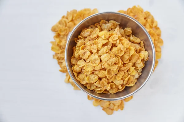 Cornflakes in a metal bowl on a painted white wooden background. The symbol of the heart is laid out of cornflakes. — Stock Photo, Image