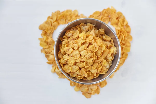 Cornflakes in einer Metallschale auf einem bemalten weißen Holzgrund. das Symbol des Herzens ist aus Cornflakes aufgebahrt. — Stockfoto