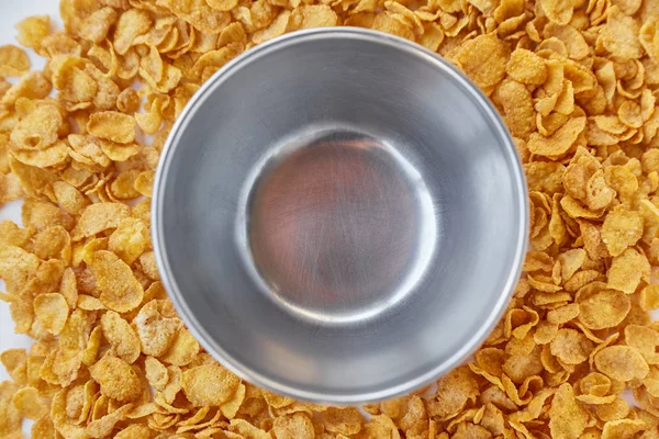 Round frame with empty metal bowl lined with corn flakes. Cornflakes scattered on a wooden table.