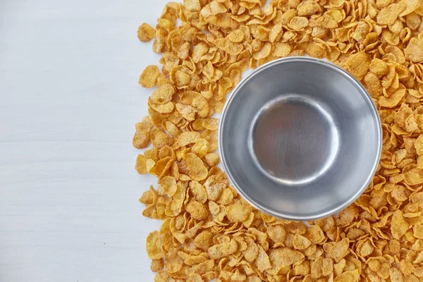 Round frame with empty metal bowl lined with corn flakes. Cornflakes scattered on a wooden table. Free space for lettering.