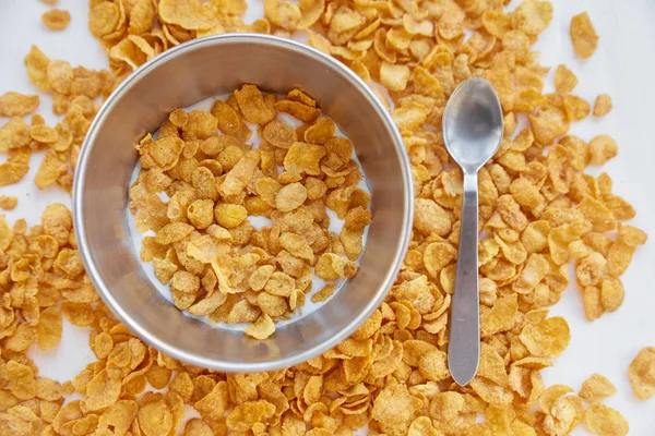 Cornflakes in einer Metallschüssel mit Milch auf einem bemalten weißen Holzgrund. Cornflakes auf Holztisch mit Löffel verstreut. — Stockfoto