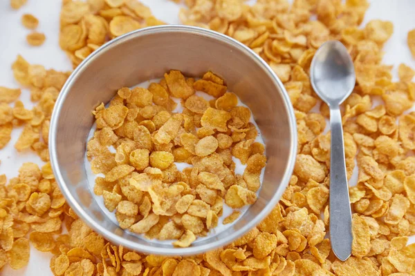 Cornflakes in einer Metallschüssel mit Milch auf einem bemalten weißen Holzgrund. Cornflakes auf Holztisch mit Löffel verstreut. — Stockfoto