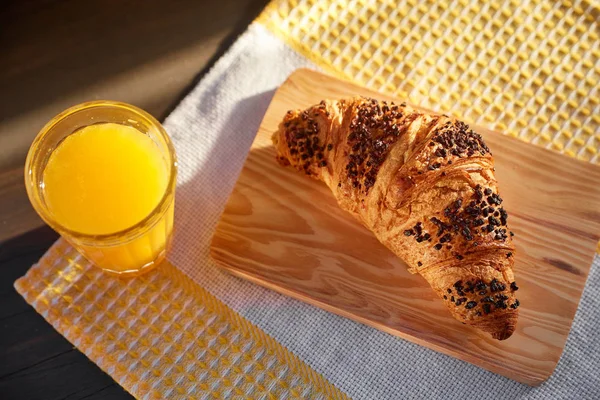 Croissant fresco em uma placa de madeira e toalha de cozinha com uma xícara de suco de laranja. Croissant recém-assado em uma mesa de madeira escura . — Fotografia de Stock